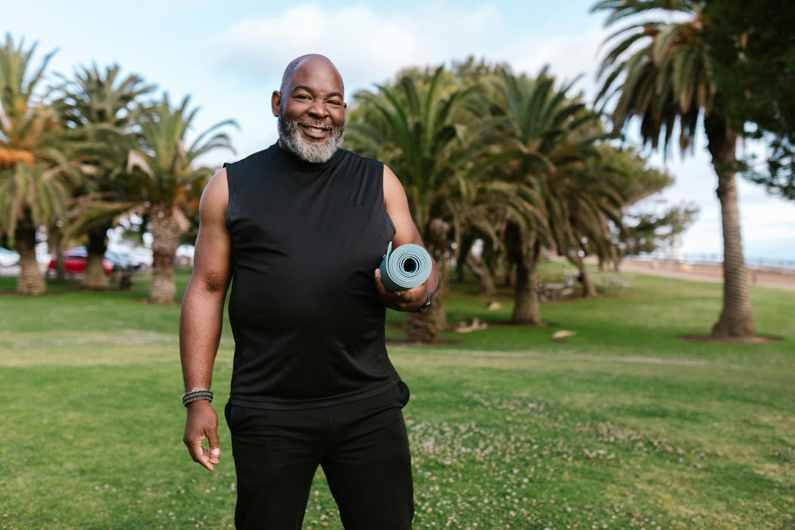Smiling elderly man holding a yoga mat in a sunny park surrounded by palm trees.