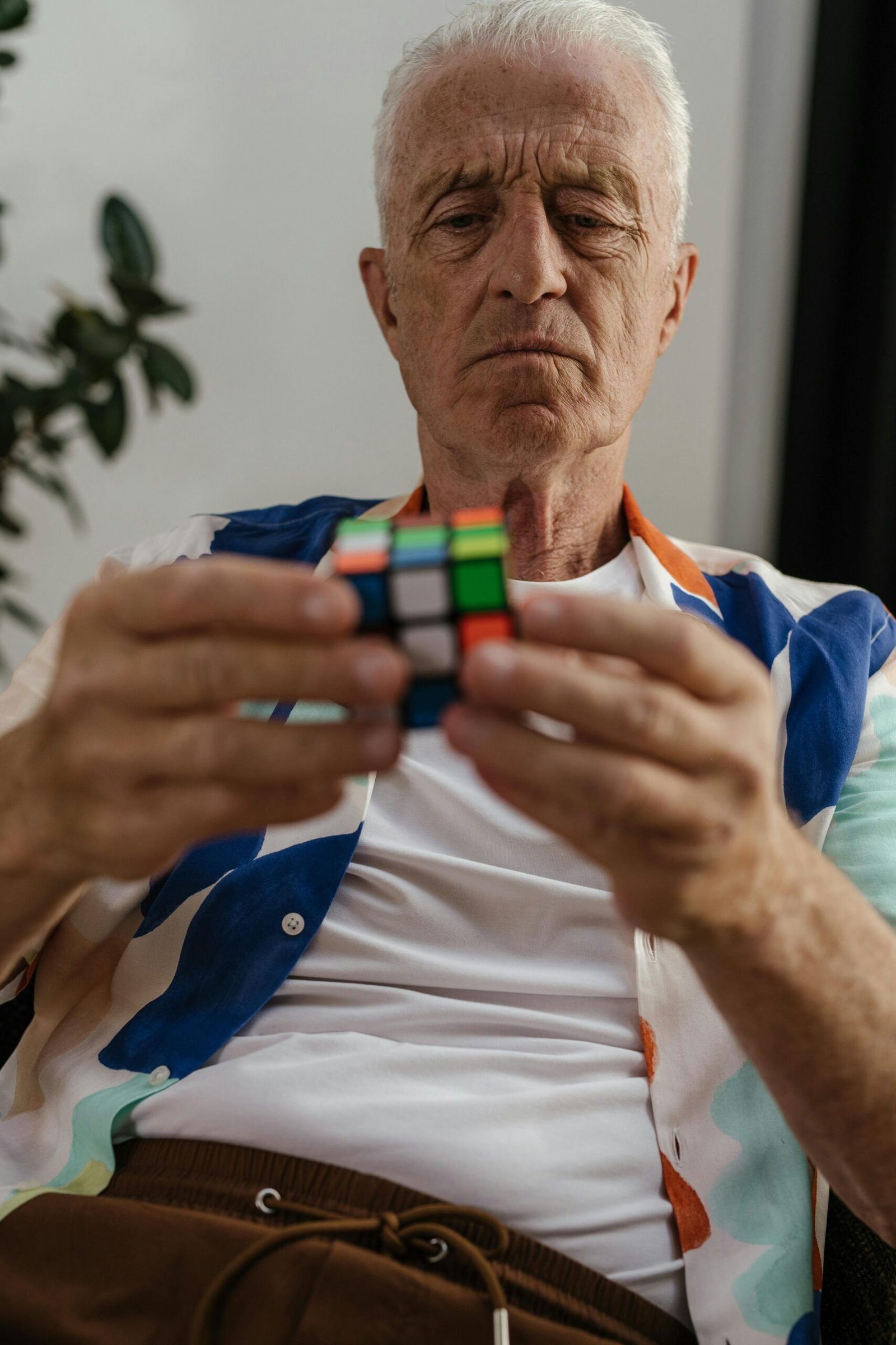 Elderly man focusing on solving a Rubik's Cube indoors, demonstrating concentration and puzzle-solving skills.