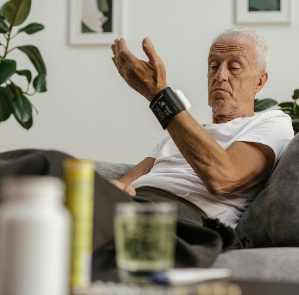 Elderly man checks his blood pressure using a wrist monitor in a comfortable living room setting.